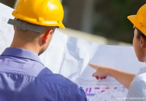 Zwei Handwerker schütteln sich die Hände über einem Tisch, auf dem zwei Helme und Pläne liegen