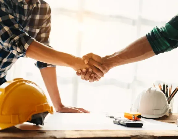 Zwei Arbeiter geben sich die Hand. Unter Ihnen ein Tisch mit Bauhelmen und Bauplan